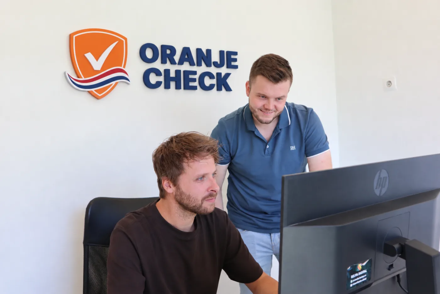 Noah en Vincent aan het werk op kantoor, onder het Oranje Check-logo aan de muur
