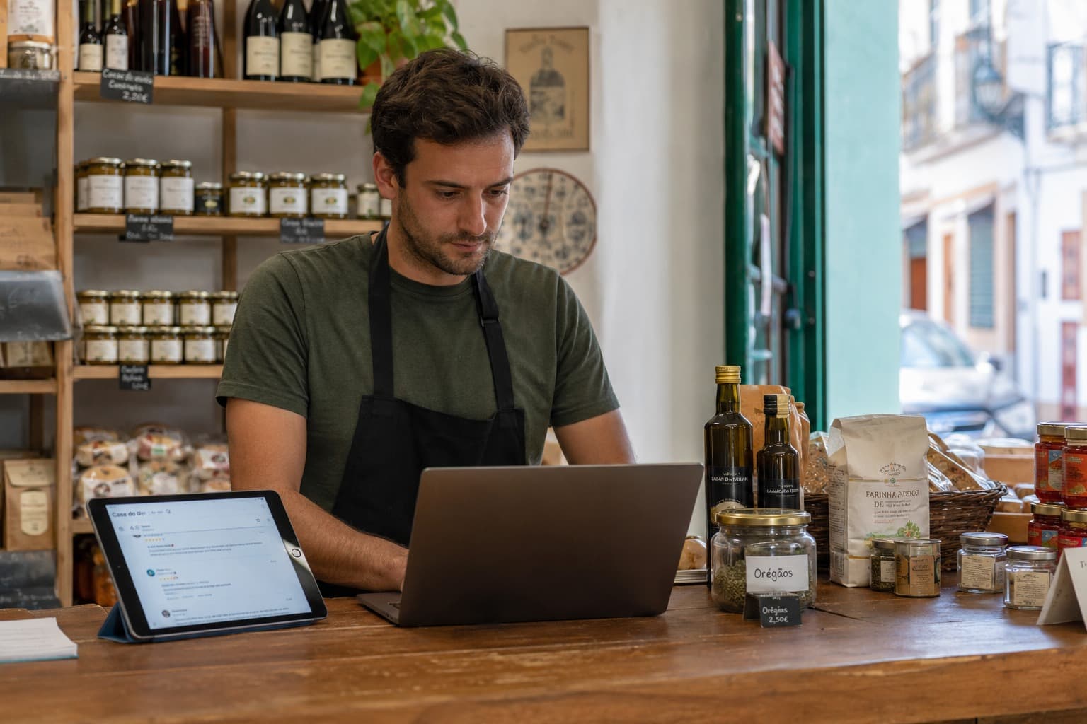 Winkelier achter de toonbank kijkt op een laptop, met naast zich een tablet waarop online recensies staan