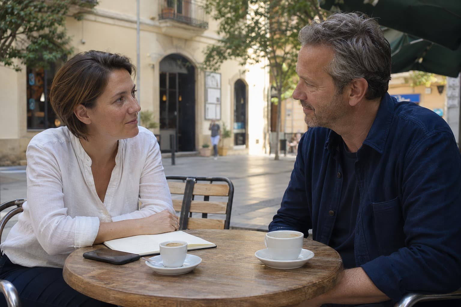 Twee mensen in gesprek aan een cafétafel op een plein in Spanje, met een opengeslagen notitieboek tussen hen in